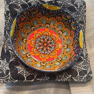 May include: A colorful bowl with an intricate floral design in shades of orange, red, and yellow sits on a black square pot holder with a white spiderweb pattern. The bowl has a blue rim.
