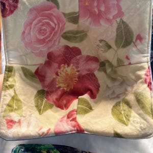 May include: A floral bowl cozy featuring a cream-colored background with large pink and red rose patterns and green leaves. The cozy is quilted with a blue trim and is designed to hold a bowl of food.