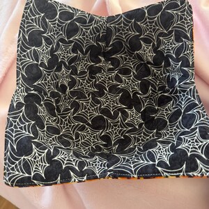 May include: A black bowl cozy with a white spiderweb pattern. The cozy is lined with orange fabric and is designed to hold a bowl of food. The cozy is made of fabric and is designed to keep food warm.