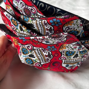 May include: Stack of red fabric bowl cozies with a sugar skull design. The cozies have a red background with white sugar skulls, blue, yellow, and purple flowers. The interior is black with a white swirl pattern.