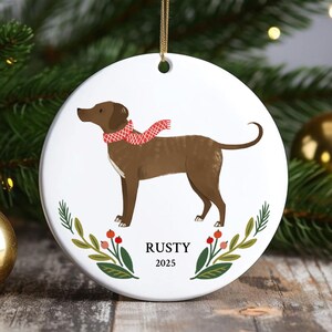 May include: A white ceramic Christmas ornament featuring a brown dog wearing a red scarf. The dog is surrounded by a floral design with the name "RUSTY" and the year "2025" printed below. A gold string is attached for hanging.