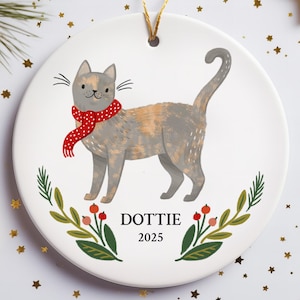 Pode incluir: Enfeite de Natal redondo de cerâmica com um gato cinzento usando um cachecol vermelho. O gato está sobre um fundo branco com detalhes florais verdes e vermelhos. O nome "Dottie" e o ano "2025" estão impressos abaixo.
