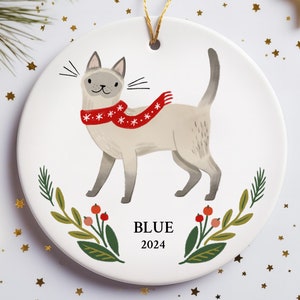May include: A white ceramic Christmas ornament featuring a cartoon Siamese cat wearing a red and white scarf. The ornament has green leaves and red berries around the cat. The text "BLUE 2024" is printed below the cat.