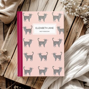 May include: A pink notebook with a burgundy spine and a repeating pattern of gray cats. The notebook cover reads "ELIZABETH JANE NOTEBOOK".