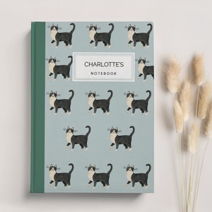May include: A notebook with a light teal cover patterned with black and white cats. The spine is green. The notebook has the text "CHARLOTTE'S NOTEBOOK" in a white rectangle. Dried beige flowers are to the right of the notebook.