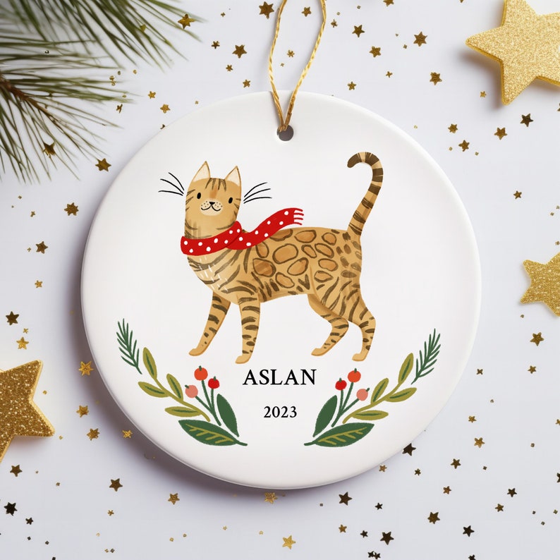 May include: A white ceramic Christmas ornament featuring a cartoon illustration of a brown tabby cat wearing a red and white polka dot scarf. The ornament has a green leafy garland with red berries and the text "ASLAN 2023".