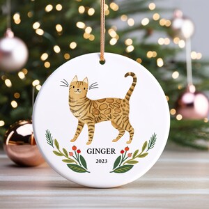 May include: A white ceramic Christmas ornament featuring a cartoon illustration of a brown tabby cat with green eyes. The cat is surrounded by green leaves and red berries. The ornament is personalized with the name "Ginger" and the year "2023".