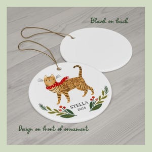 May include: A white ceramic Christmas ornament with a brown tabby cat wearing a red scarf and a green and red floral design. The ornament is personalized with the name "STELLA" and the year "2024".