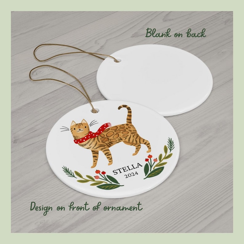 May include: A white ceramic Christmas ornament with a brown tabby cat wearing a red scarf and a green and red floral design. The ornament is personalized with the name "STELLA" and the year "2024".