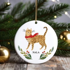 May include: White ceramic Christmas ornament featuring a cartoon illustration of a brown tabby cat wearing a red and white polka dot scarf. The ornament has green leaves and red berries around the cat and the name "NALA" printed below the cat.