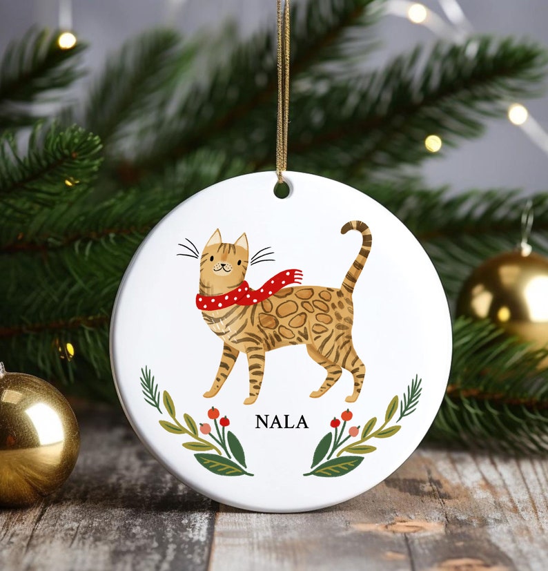 May include: White ceramic Christmas ornament featuring a cartoon illustration of a brown tabby cat wearing a red and white polka dot scarf. The ornament has green leaves and red berries around the cat and the name "NALA" printed below the cat.