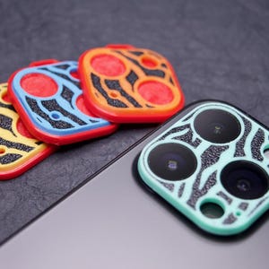May include: Three colorful camera lens covers with a zebra print design and a green camera lens cover with a zebra print design on a black smartphone.