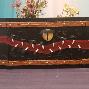 May include: A black wooden box with a red and white painted design of a monster's mouth with a yellow eye. The box has a brown leather-like trim with gold studs.