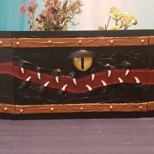 May include: A black wooden box with a red and white painted design of a monster's mouth with a yellow eye. The box has a brown leather-like trim with gold studs.