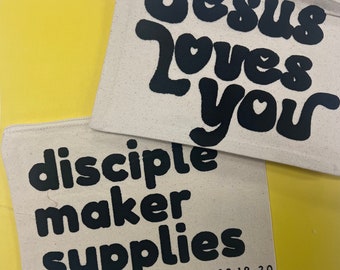 May include: Two white canvas zipper pouches with black text. The top pouch has the text "Jesus Loves You" in a bubble font. The bottom pouch has the text "disciple maker supplies" in a bubble font and the text "MATTHEW 28:19-20" in a smaller font.
