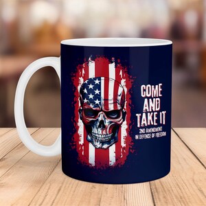 May include: A white ceramic coffee mug with a blue background and a skull design. The skull is wearing a bandana with the American flag design. The text "Come and Take It" is printed on the mug, along with "2nd Amendment in Defence of Freedom".