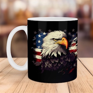 May include: A white ceramic mug with a black handle features a graphic of a bald eagle with a close-up view of its head and beak. The eagle is positioned in front of a faded American flag with red, white, and blue stripes and stars.