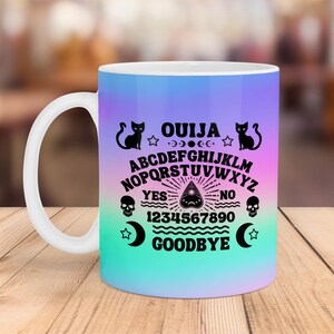 May include: A white ceramic mug with a colorful gradient design featuring a black and white Ouija board with the text "OUIJA", "YES", "NO", "GOODBYE", and numbers 1-9 and 0. The design also includes black cats and stars.