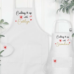 Puede incluir: Dos delantales blancos con corazones rojos y el texto "Cooking it up with" seguido de un nombre en letras doradas. El delantal de la izquierda dice "Caitlyn" y el delantal de la derecha dice "Grandma".
