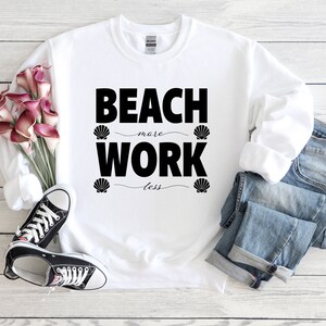 Puede incluir: Sudadera blanca con texto negro que dice "Beach more, Work less" con un gráfico de concha en cada lado del texto.