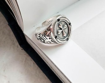 May include: Silver ring with a black cross design and black stones. The ring has a detailed floral pattern on the band. The ring is marked with the number "925".