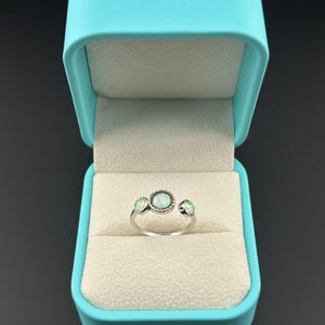 May include: A silver ring with three opal stones, presented in a turquoise jewelry box. The ring features a central oval opal stone, flanked by two smaller round opals. The box has a soft, cream-colored interior.