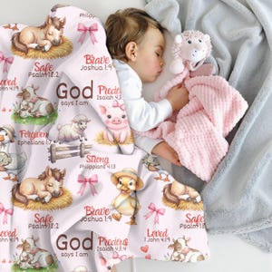 May include: A baby sleeps under a pink and white blanket with illustrations of farm animals and text. The blanket features a horse, cow, pig, sheep, and duckling, with phrases like "Safe" and "Loved". A pink plush toy and a gray blanket are also visible.