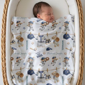 May include: A white baby blanket with a baseball theme, featuring illustrations of teddy bears in baseball uniforms, baseballs, and baseball equipment. The blanket has the text "Michael Joseph" and "Born to play BALL".