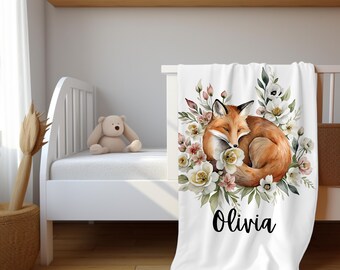 May include: White baby blanket featuring a watercolor illustration of a sleeping fox surrounded by flowers. The fox is orange and brown, and the flowers are cream, pink, and green. The name "Olivia" is written in black script.