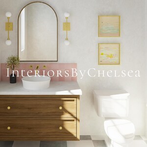 May include: A bathroom vanity with a wooden cabinet, two drawers with gold knobs, a white countertop, and a round vessel sink. Above, an arched mirror is flanked by two gold wall sconces. Pink tiles line the wall, and two framed art pieces hang nearby.