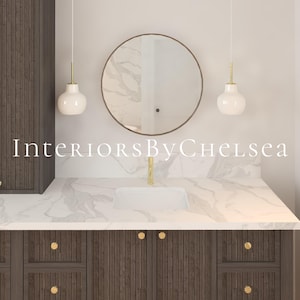 May include: A bathroom vanity with a white marble countertop and dark wood cabinets. The vanity has a round mirror with a gold frame and two white pendant lights with gold accents. The text "Interiors By Chelsea" is visible on the countertop.