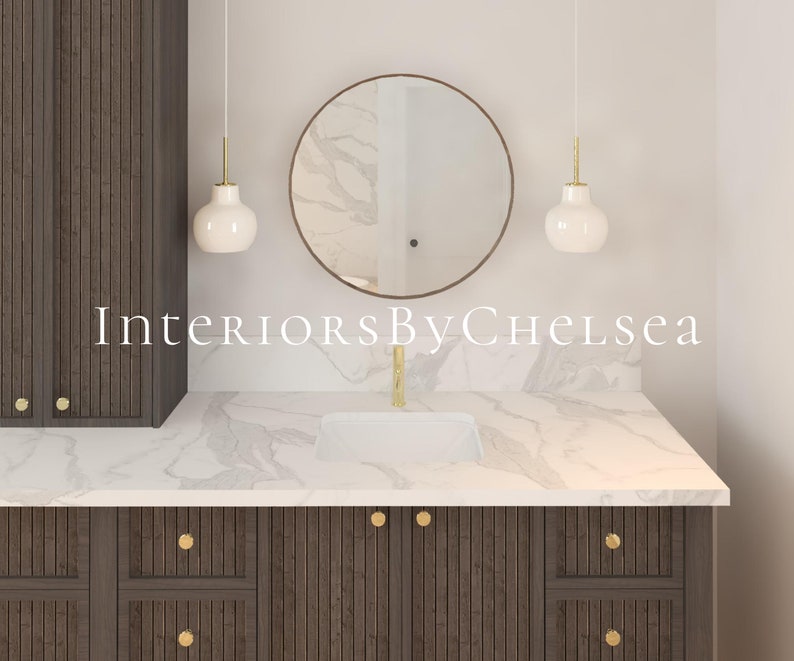 May include: A bathroom vanity with a white marble countertop and dark wood cabinets. The vanity has a round mirror with a gold frame and two white pendant lights with gold accents. The text "Interiors By Chelsea" is visible on the countertop.
