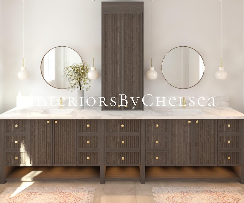 May include: A modern bathroom vanity with a white marble countertop and dark wood cabinets. The vanity has two round mirrors, two white pendant lights, and gold hardware. The text "Interiors By Chelsea" is visible on the countertop.