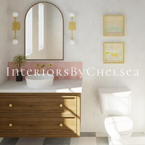 May include: A white bathroom with a wooden vanity with two drawers, a white porcelain sink, a white toilet, and a pink tile backsplash. The vanity has gold knobs. There is an arched mirror above the sink with two gold sconces on either side. There are two framed abstract paintings on the wall above the toilet.