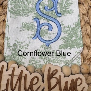 May include: A white fabric towel featuring a green toile pattern of figures and trees, embroidered with a large cornflower blue letter S. The text Cornflower Blue is printed above a wooden sign reading Little Blue Heron Embroidery.