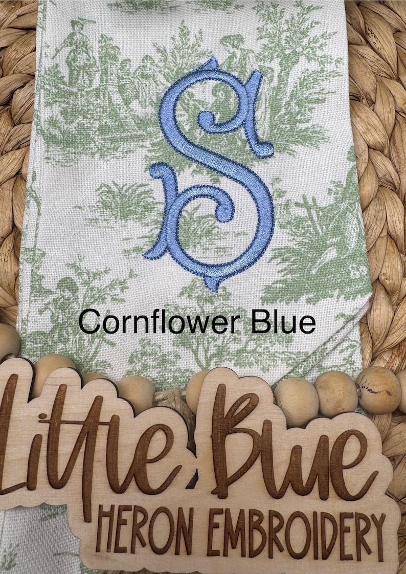 May include: A white fabric towel featuring a green toile pattern of figures and trees, embroidered with a large cornflower blue letter S. The text Cornflower Blue is printed above a wooden sign reading Little Blue Heron Embroidery.