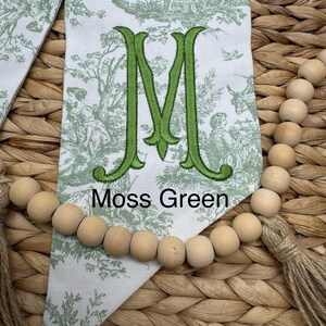 May include: A white fabric pennant featuring a green toile pattern of figures and trees, centered with a large embroidered green letter M and the text Moss Green, accented by a string of natural wooden beads and a jute tassel at the bottom.