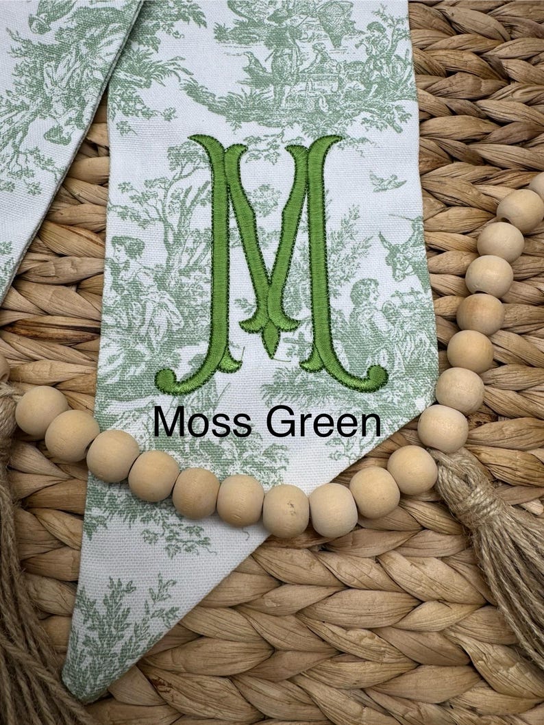 May include: A white fabric pennant featuring a green toile pattern of figures and trees, centered with a large embroidered green letter M and the text Moss Green, accented by a string of natural wooden beads and a jute tassel at the bottom.