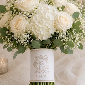 May include: A floral bouquet featuring white roses, white hydrangea, and baby's breath with green eucalyptus leaves. The stems are wrapped in a white fabric band embroidered with a gold monogram of the letters C and K and the date 9.26.26.