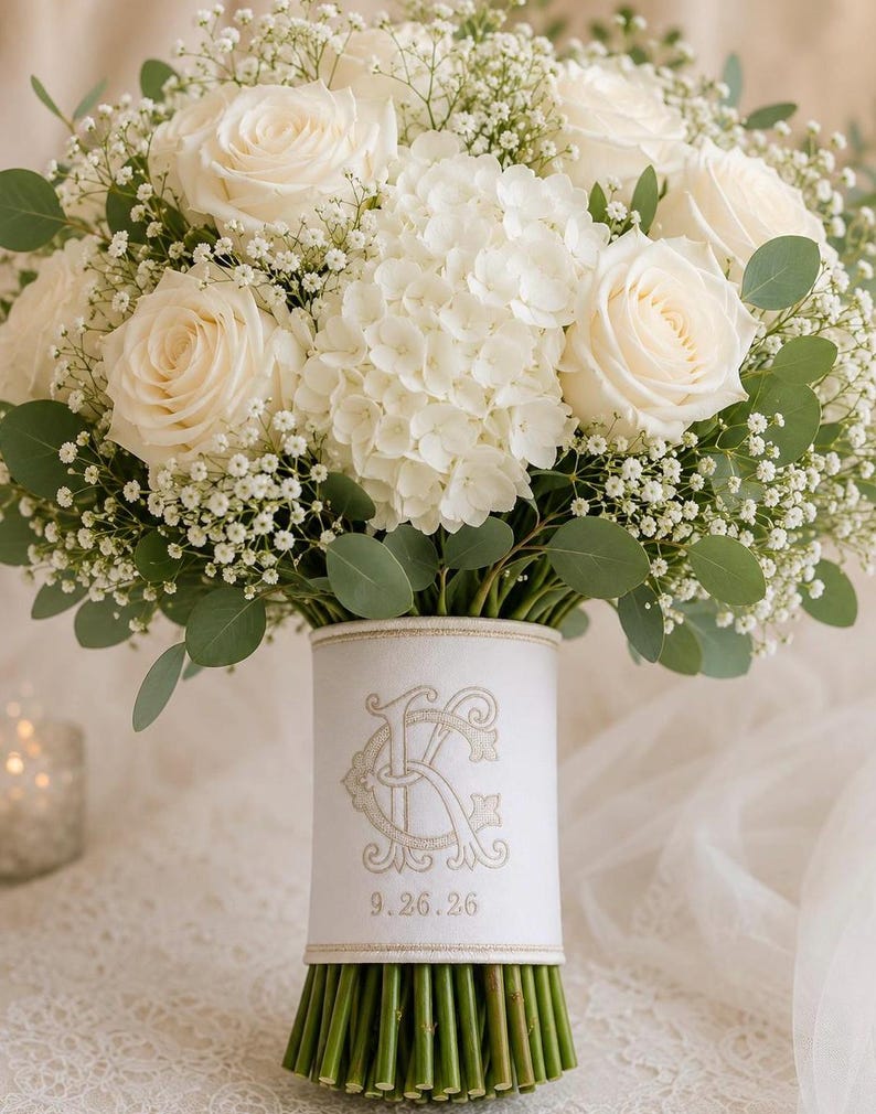 K&ouml;nnte beinhalten: Ein Blumenstrau&szlig; aus wei&szlig;en Rosen, wei&szlig;en Hortensien und Schleierkraut mit gr&uuml;nen Eukalyptusbl&auml;ttern. Die Stiele sind mit einem wei&szlig;en Stoffband umwickelt, das mit einem goldenen Monogramm der Buchstaben C und K sowie dem Datum 9.26.26 bestickt ist.
