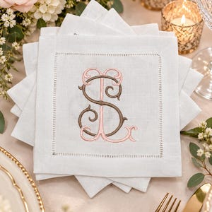 May include: A stack of square white linen napkins featuring a hemstitched border and a central embroidered monogram. The monogram consists of the letters S and L intertwined, with the letter S in brown and the letter L in light pink thread.