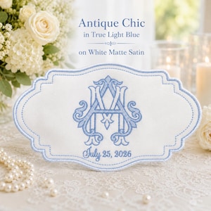 K&ouml;nnte beinhalten: Eine wei&szlig;e, matte Satin-Stoffplakette mit gewelltem Rand, die eine hellblaue Stickerei des Monogramms AM und das Datum July 25, 2026 zeigt. Der Text Antique Chic in True Light Blue on White Matte Satin steht &uuml;ber der Plakette.