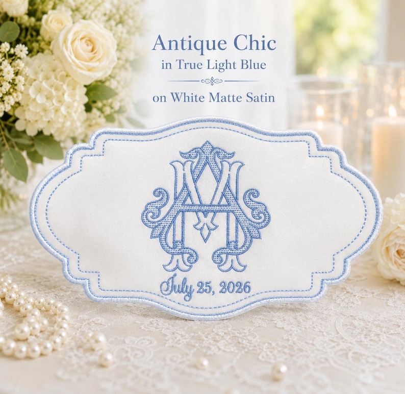 K&ouml;nnte beinhalten: Eine wei&szlig;e, matte Satin-Stoffplakette mit gewelltem Rand, die eine hellblaue Stickerei des Monogramms AM und das Datum July 25, 2026 zeigt. Der Text Antique Chic in True Light Blue on White Matte Satin steht &uuml;ber der Plakette.