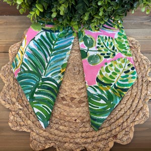 May include: Two pink fabric squares with a green and white tropical leaf pattern. The fabric squares are on a woven straw placemat.
