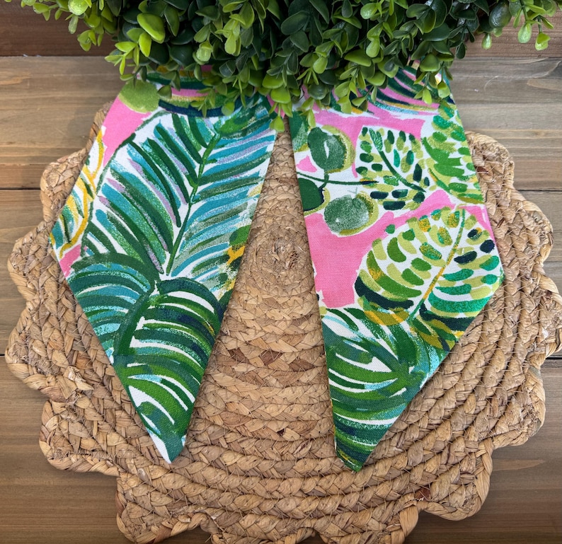 May include: Two pink fabric squares with a green and white tropical leaf pattern. The fabric squares are on a woven straw placemat.