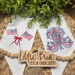 May include: Two white fabric pennants featuring machine embroidery. The left pennant displays two crossed American flags tied with a red bow. The right pennant features a large, ornate, red and blue embroidered monogram letter R. A wooden sign reads Little Blue Heron Embroidery.