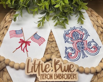 May include: Two white fabric pennants featuring machine embroidery. The left pennant displays two crossed American flags tied with a red bow. The right pennant features a large, ornate, red and blue embroidered monogram letter R. A wooden sign reads Little Blue Heron Embroidery.