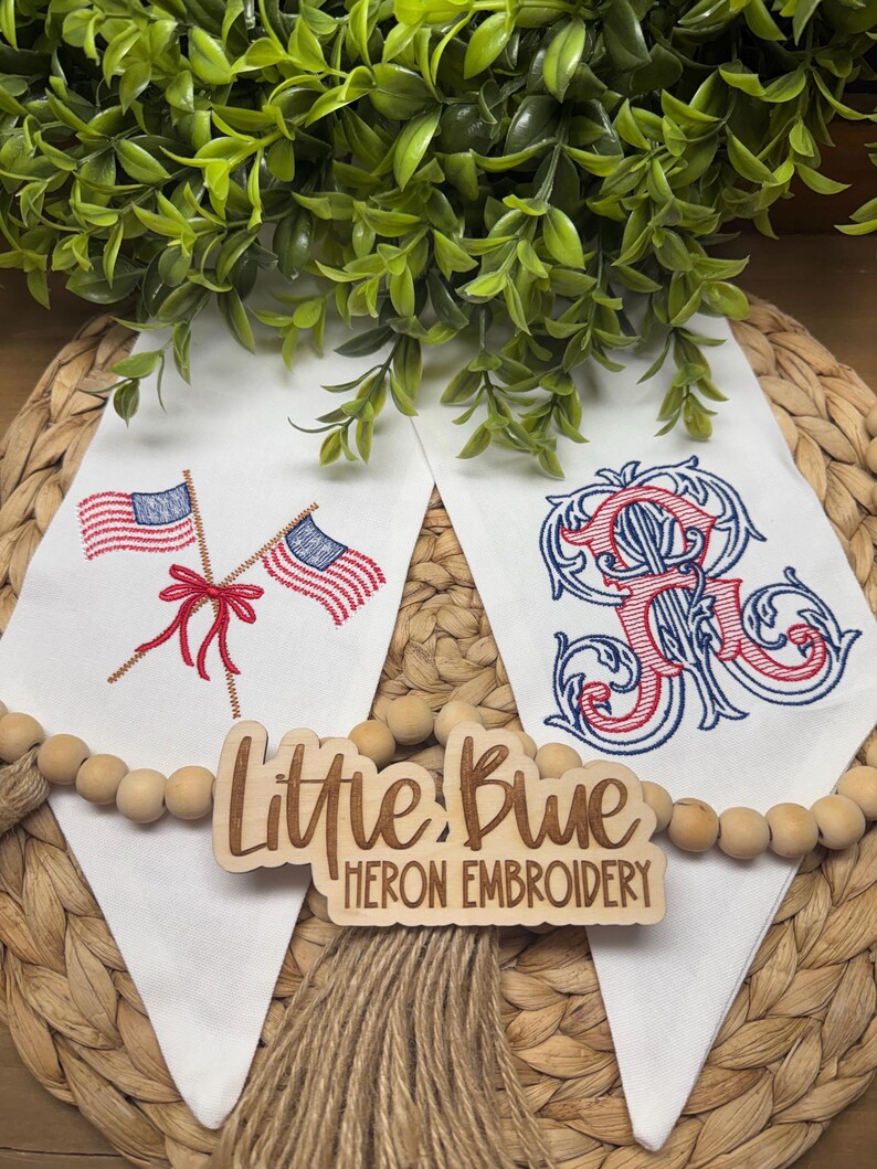 May include: Two white fabric pennants featuring machine embroidery. The left pennant displays two crossed American flags tied with a red bow. The right pennant features a large, ornate, red and blue embroidered monogram letter R. A wooden sign reads Little Blue Heron Embroidery.