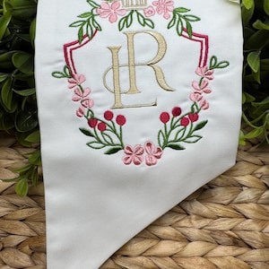May include: A white fabric item featuring an embroidered design of the letters LR in gold thread, centered within a floral wreath of pink flowers, green leaves, and red berries, topped with a small gold architectural icon in a shield shape.