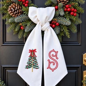 Peut inclure: Un nœud en tissu blanc suspendu à une couronne composée d'aiguilles de pin vertes, de pommes de pin brunes et de baies rouges. Le pan gauche présente un sapin de Noël brodé avec un nœud rouge, et le pan droit porte une lettre S rouge.